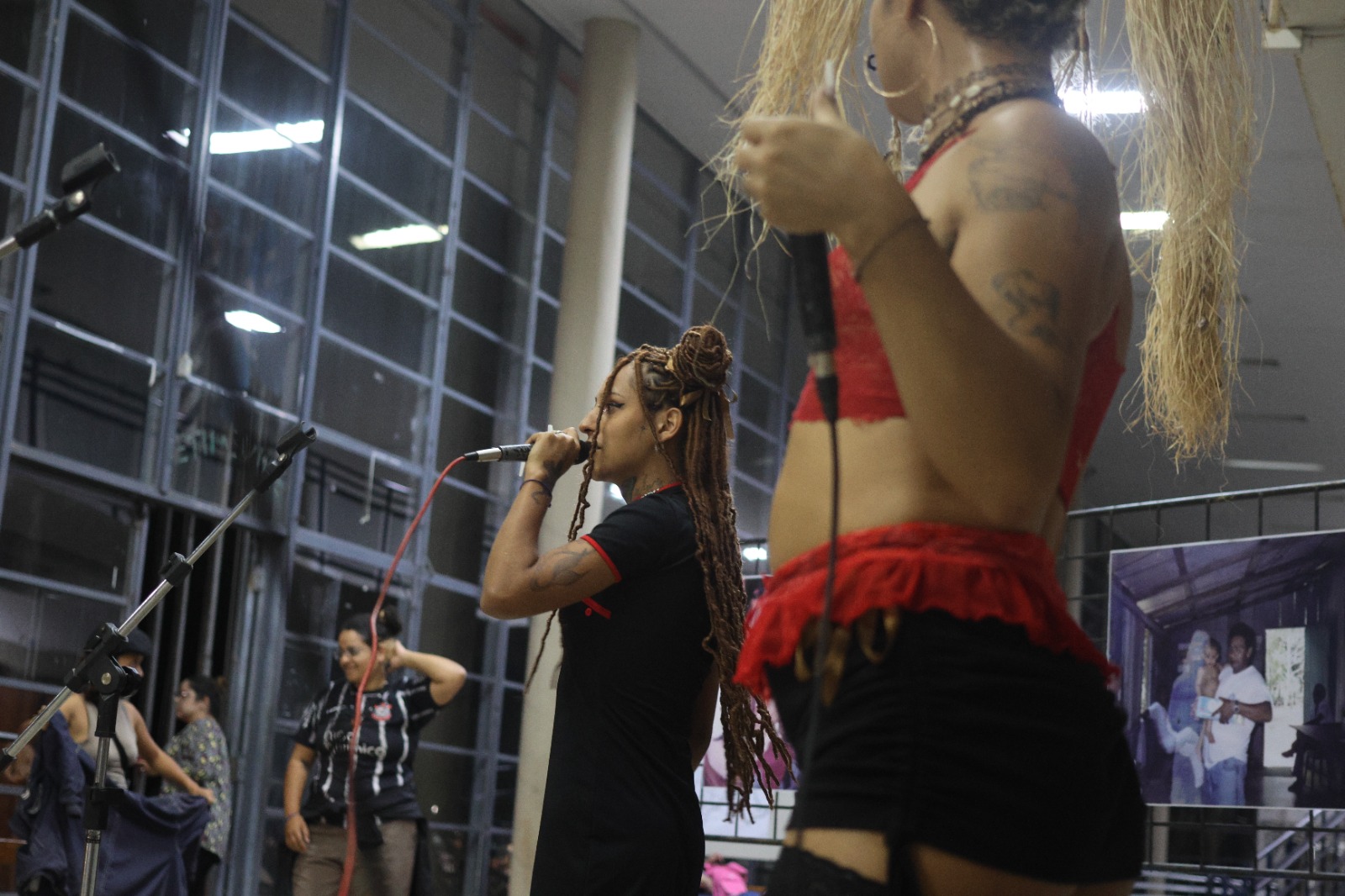  Fotografia. Em destaque, duas pessoas ao microfone. A da esquerda é alta, negra de pele retinta, tem tranças no cabelo e usa um penteado de dois coques altos. Usa um vestido preto. A pessoa da direita é baixa, negra e de pele clara, tem os cabelos curtos e cacheados. Ela usa boné preto, camiseta cinza-escuro e bermuda azul. As duas pessoas seguram microfones de fios nas mãos. O espaço tem dois murais com cartazes: o da esquerda estampa uma foto de um homem com um bebê no colo e uma silhueta em sombra clara à direita dele. No cartaz da direita, há o título em letras pretas: “O passado que nunca passa”. Presas ao teto, duas instalações de corda pretas com nós e palha em cascata.  
