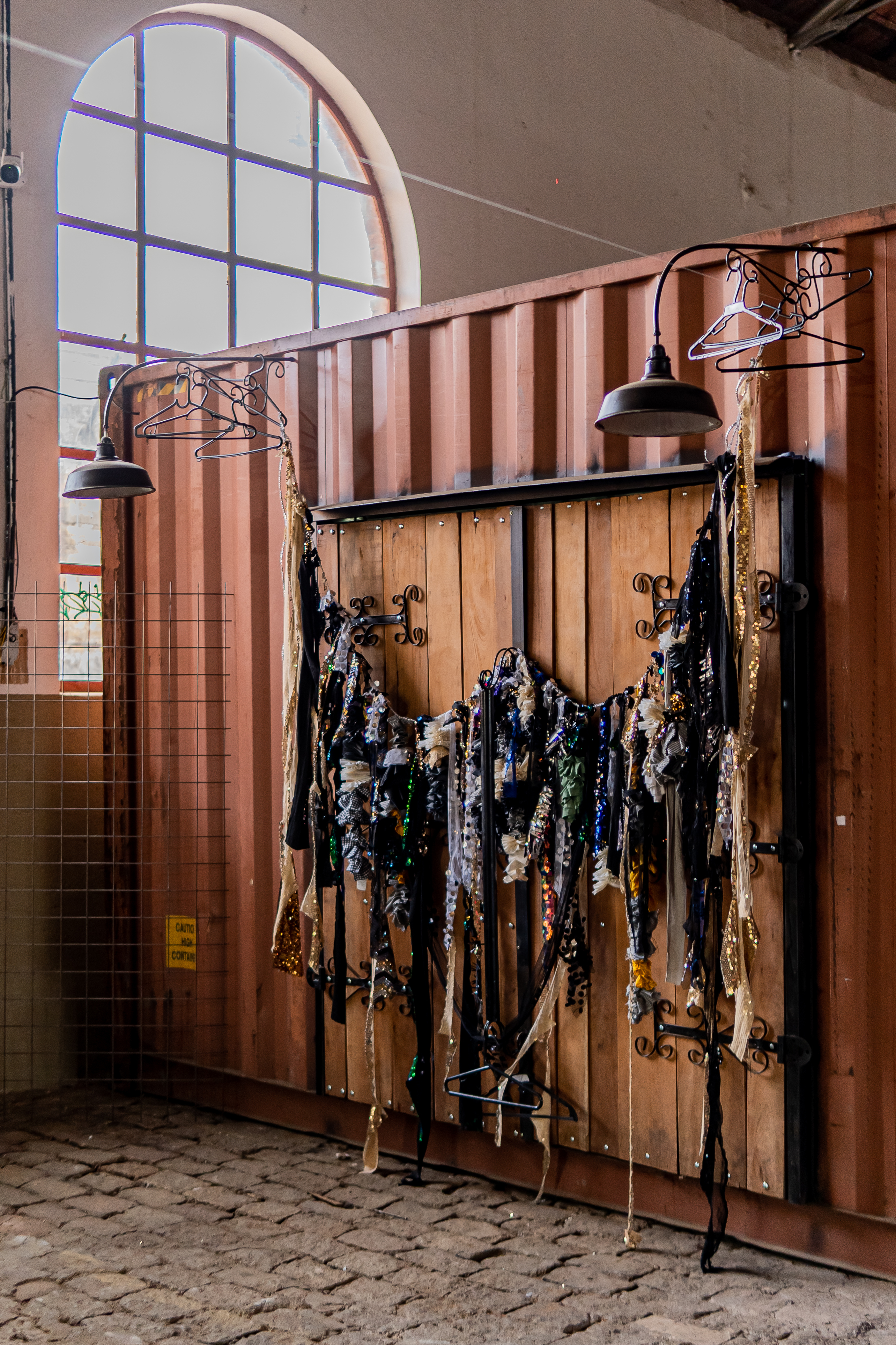 Fotografia de uma instalação. Em destaque, uma porta de madeira com ganchos ornados, pendurado na horizontal e rente a porta de madeira um fio com diversas tiras e tecidos coloridos, com brilhos, lantejoulas e fitas. 
Ao fundo, uma parede de ferro marrom, na parte superior duas arandelas de poste antigas e na haste diversos cabides.  
