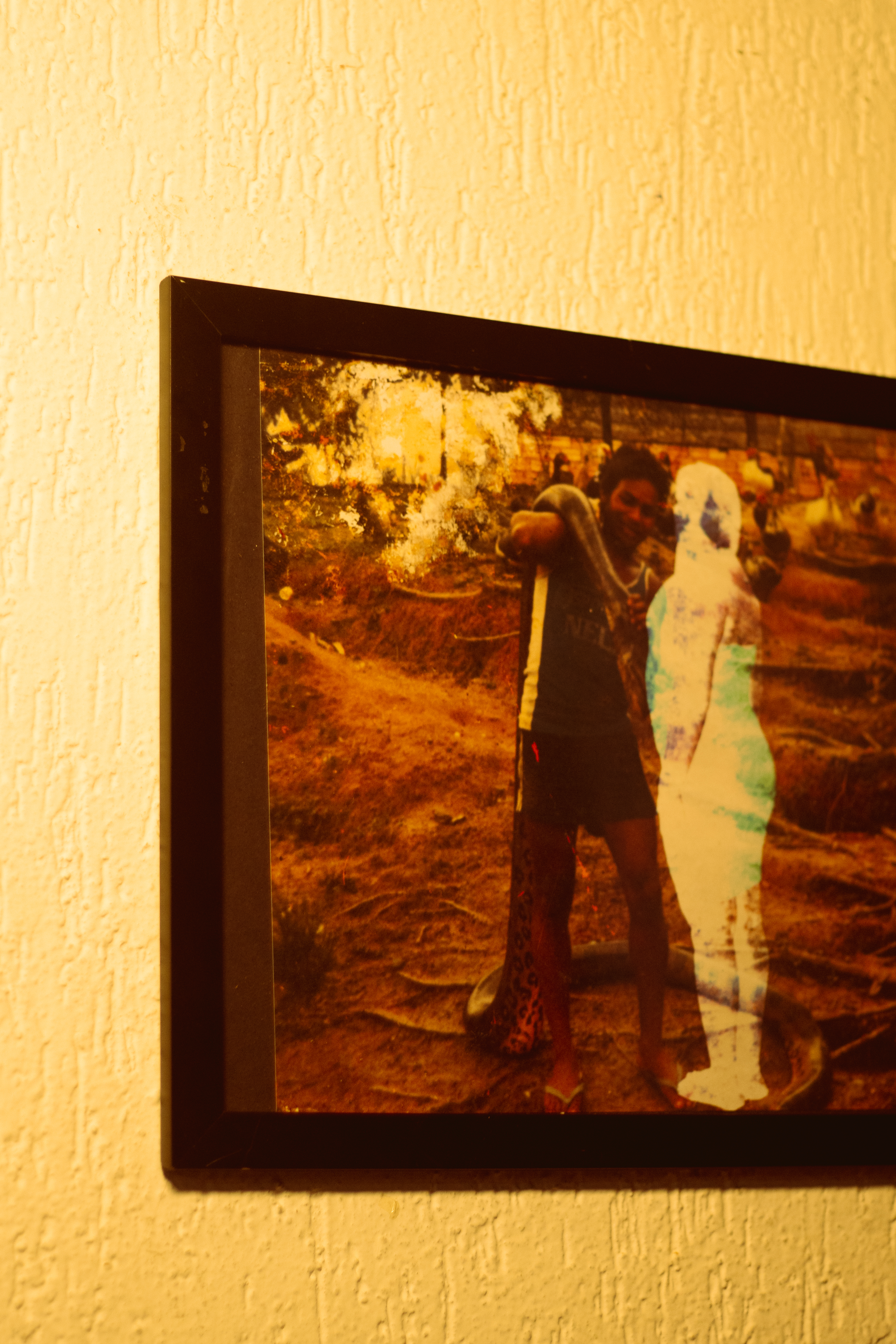 Fotografia de um quadro com foto. Ambiente rural, com chão de terra batida e raízes expostas. Um homem de pele escura, vestindo camiseta regata escura, shorts e chinelos, está de pé segurando uma cobra de grande porte enrolada ao redor de seu corpo. Ele olha para a câmera com expressão neutra. Em frente a ele, há a silhueta de uma figura feminina translúcida, com coloração azulada e esbranquiçada, tratada digitalmente, contrastando com o estilo fotográfico antigo e desbotado da imagem. Ao fundo, há galinhas espalhadas pelo terreno. 

